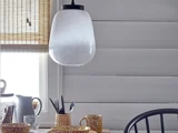 En pendel i matteret glas hænger over et spisebord med plettede kopper, en kande, bestik og en kurvholder. Baggrunden har hvide træpaneler og et vindue med en bambusskærm.