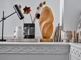 En hvid kaminhylde viser moderne vaser, abstrakte skulpturer, en sort skrivebordslampe, tørrede blomster, bøger og et lyserødt stearinlys i en struktureret, minimalistisk indretning.