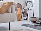 Et moderne stuehjørne med abstrakt kunst, en solbrun skulptur, indrammede tryk, en keramikskål og neutral indretning på et hvidt gulv ved siden af en beige sofa.