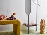 En gul bænk med en stribet pude, keramikvase og bakke står ved siden af et moderne spejlstativ med en bog; en abstrakt træskulptur står på gulvet ved en hvid væg.
