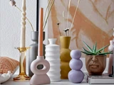 En samling farverige moderne vaser, unikke lysestager og en aloeplante i en ansigtsformet potte, arrangeret på en flade foran abstrakt vægkunst.