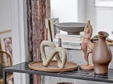 En keramisk bakke rummer to moderne, sløjfeformede lysestager og stearinlys. I nærheden står kunstbøger, en grå skål, en solbrun kande og en lille abstrakt menneskeskulptur på en sort-hvid hylde.