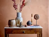 Et træbord med keramiske vaser, tørrede blomster, et stearinlys, en pyntegenstand, et rundt keramikstykke og bøger, der står foran en ferskenfarvet væg.