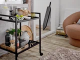 En sort barvogn rummer drinks, glas og en skål med limefrugter i et hyggeligt opholdsrum med en brun stol, et lurvet tæppe og bøger stablet nær en døråbning.