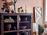 Et træskab med glasdøre viser dekorative skåle og vaser. På toppen står lysestager, små planter og en kande. Kunst og en lampe står ved siden af en seng med strukturerede puder.