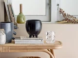 Et rattanbord med dekorative vaser, et glas med boblelignende design, bøger, tørrede blade og et stearinlys foran et hvidt vindue.