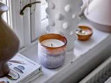 Et stearinlys i en gradientbrun og hvid holder står i vindueskarmen ved siden af en hvid, struktureret vase, en lille skål og to bøger ved et vindue.