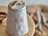 En keramikkrukke mærket TREATS med en knogleformet hank på låget, der står på en træplade med et sløret hundehalsbånd i baggrunden.