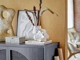 En moderne skænk med abstrakt indretning, herunder en skulpturel vase, tørrede grene, kunsttryk, en stribet kasse og stablede bøger mod en solbrun væg.