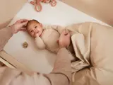 En baby i beige tøj ligger i en krybbe, delvist dækket af et tæppe, mens en voksen forsigtigt rører ved babyens hoved og holder hånden. En sut og et tøjdyr er i nærheden.