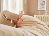 Nærbillede af en babys fødder iført beige strikbukser, der hviler på et blødt cremefarvet tæppe i et blødt oplyst, hyggeligt rum.