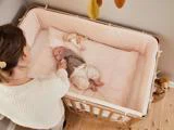 En kvinde står ved siden af en krybbe af træ og holder forsigtigt en baby i hånden, som ligger på en blød, lyserød madras med et tøjdyr i nærheden.