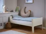 En lille, hvid børneseng med blåt sengetøj, en halvmånepude og en løve som legetøj i et hyggeligt rum med bøger, en lampe og en flettet kurv på et trægulv.