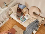 Et lille barn sidder på en seng og kigger i en opslået billedbog. Sengen har beige sengetøj, en halvmåneformet pude og et blåt tæppe. Trægulv og legetøj er synligt i nærheden.