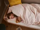 Et lille barn sover i en seng og kæler for et tøjdyr under et lyserødt tæppe med en gul pude og en bog på gulvet i nærheden.