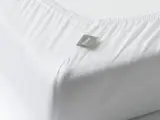 Nærbillede af et hvidt lagen med elastiske kanter på en madras, med et lille gråt mærke med påskriften FLEXA.