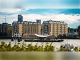 En moderne bygning ved floden står bag en lille dok på vandet, med bybygninger og grønne områder i baggrunden under en delvis overskyet himmel.