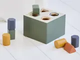 Et formsorteringslegetøj med en grøn kasse og forskellige farverige træfigurer, bl.a. cylindre, sekskanter og en trekant, på en hvid overflade.