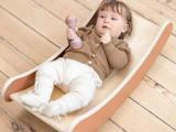 En baby ligger på et buet balancebræt af træ, holder en lille rangle og er iført en brun sweater, hvide bukser og bløde sko på et trægulv.