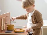 Et lille barn leger med et legetøjskøkken og lader, som om det laver mad med en gryde og en pande med legetøjsmad, mens det er iført en brun sweater og en hvid skjorte.