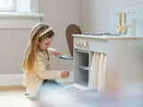 En ung pige leger med et legetøjskøkken og sætter en lille gryde med legetøjsmad ind i en legetøjsovn i et svagt oplyst, hyggeligt rum.