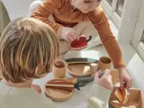 To børn leger med legetøjsmad og -redskaber af træ på et hvidt bord og lader, som om de skærer og serverer stykker i et lyst, solbelyst rum.