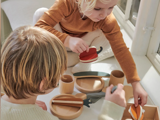 To små børn leger med legetøjsmad og -tallerkener af træ på en hvid flade nær et vindue, fokuseret og engageret i en fælles leg.