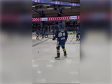 Ishockeyspillere i blå trøjer varmer op på isbanen, og i forgrunden står en spiller med en stav og en puck.