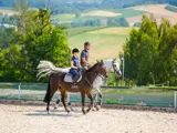 To personer med hjelme rider på heste side om side i en udendørs arena med et naturskønt landskab af marker, træer og bakker i baggrunden.