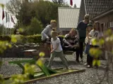 En ung pige spiller minigolf, mens fire personer ser på og smiler bag hende på en solrig dag med bygninger og grønne områder i baggrunden.
