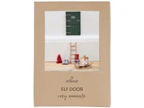 En grøn miniaturedør, stige, rød hat, støvler, lanterne og kælk er arrangeret mod en hvid væg på en træoverflade, udstillet på en brun emballage mærket Elf Door cosy moments.