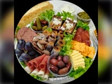 Et fad med forskellige oste, kød, vindruer, oliven, kiks, tørret frugt, nødder, brownies, pickles, brød, dip og salat, der er arrangeret pænt til servering.