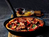 En stegepande fyldt med en velsmagende pandekage toppet med bacon, tomatskiver og krydderurter, anbragt på et træbræt med sennep, marmelade og brødskiver i baggrunden.