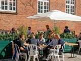 Folk sidder ved borde uden for en café med hvide stole og en stor parasol ved siden af en murstensbygning på en solrig dag. Nogle mennesker går forbi i baggrunden.