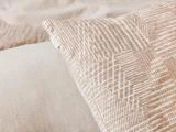 Nærbillede af en beige tekstureret pude med et geometrisk mønster, der hviler på en lys stofoverflade og antyder en hyggelig og minimal soveværelsesindretning.