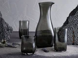 En glaskaraffel og tre røggrå glas er anbragt på en struktureret overflade med vulkanske sten og tørret tidsel i baggrunden.