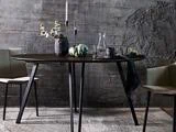 Et moderne spisebord med sorte ben, dækket med en vase med blomster, stearinlys, en glaskaraffel og artiskokker, flankeret af to beige stole mod en struktureret grå væg.
