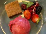 En tallerken med et firkantet stykke kage, en kugle lyserød sorbet og en pynt af friske bær og mynteblade.