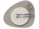 Minimalistisk borddækning med en rund beige tallerken, en lille beige skål, gaffel og kniv på en grå, asymmetrisk dækkeserviet.