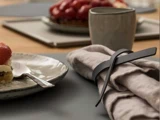 Nærbillede af en borddækning med en grå serviet omviklet med en sort læderring, en keramiktallerken med dessert og en ske samt et krus i baggrunden.