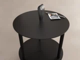 Rundt sort sidebord med en struktureret overflade, en håndtagsløkke på toppen og et lille foto og en kuglepen placeret på overfladen. Baggrunden er en neutral beige farve.