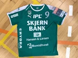 En grøn sportstrøje med hvid skrift, nummer 9 og flere sponsorer, bl.a. SKJERN BANK, ligger på et trægulv i en gymnastiksal med gule og orange banelinjer.