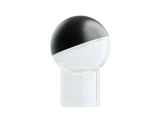 En moderne bordlampe med en rund kuppel, der er halvt sort og halvt gennemsigtig, og som står på en gennemsigtig, cylindrisk fod, set på en hvid baggrund.
