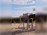 Børn leger på en sandlegeplads med gynger, rutsjebane og klatreredskaber under en delvis overskyet himmel. Et barn sidder på en gynge, et andet sidder i sandet.