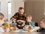 En skægget mand og tre børn sidder ved et spisebord og deler et måltid med drikkevarer, brød og forskellige retter i et moderne, lyst rum.