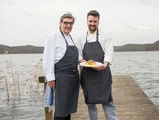 To kokke i stribede forklæder står på en træbrygge ved en sø og smiler, mens den ene holder en tallerken med mad. Baggrunden viser vand og fjerne træer under en overskyet himmel.