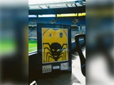 Indrammet gul fodboldtrøje med en sort edderkopgrafik på en baggrund af et fodboldstadion.