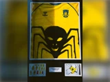 En indrammet gul fodboldtrøje med et stort sort edderkoppedesign, Brøndby IF-logo og tre små designskitser nedenunder er udstillet.