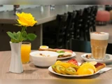 Et morgenbord med en gul rose i en vase, et glas appelsinjuice, en skål morgenmadsprodukter, en latte og en tallerken med udskåret frugt og kager.