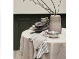 Et rundt bord med en beige dug, stablede tallerkener, kopper, servietter, et stribet viskestykke, en sølvskål og en stor beige vase med nøgne grene.